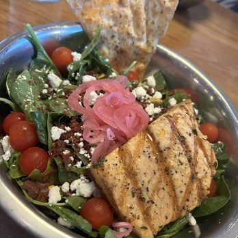 Crust Brewing - Salad with salmon. Really enough for 2 for lunch. - A salad with grilled salmon fillet, cherry tomatoes, pickled pink onions, and crumbled feta cheese is presented in a metal bowl. The salad includes leafy greens and is garnished with herbs. A slice of flatbread is placed upright in the bowl. The background shows a wooden table and a glass of water. - Breweries Near Me - Rosemont, Illinois