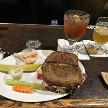 Friar Tuck's - Corned beef. - A plate contains a sandwich made with dark bread, with visible meats and cheese, along with pickles, a small container of sauce, and a slice of carrot. Two drinks are placed behind the plate: one red-colored cocktail with ice and garnishes, and one yellowish beverage with ice. The setting appears to be a bar or restaurant with a dark wooden counter. - American Near Me - Manitowoc, Wisconsin
