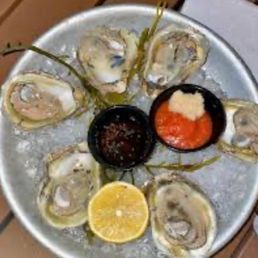 Happy hour oysters for $1 - Six raw oysters on the half shell are arranged on a metal tray, accompanied by lemon slices and two small containers of cocktail sauce and mignonette.
