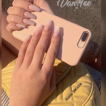 Vanitee Spa - Two hands with well-maintained, natural-colored manicured nails are holding a smartphone with a pink case. The background includes a yellow quilted bag with a zipper and a metallic logo, and the setting appears to be outdoors with a textured ground surface. The image emphasizes the nails and the phone, with a logo reading 'Vanitee Nails' in the upper right corner. - Nail Salons Near Me - Lansing, Michigan