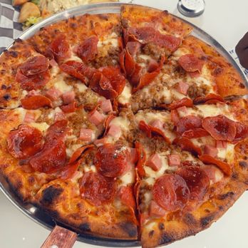 The Station - The station pizza - A pizza with a golden-brown crust topped with pepperoni, ham, sausage, and melted cheese is presented on a round metal tray with a wooden handle. The pizza is cut into slices, and the toppings are evenly distributed. In the background, a person wearing a black and pink jacket is seated at a table with a plate of rice and vegetables. The setting appears to be a casual dining environment. - Pizza Near Me - Jackson, Mississippi