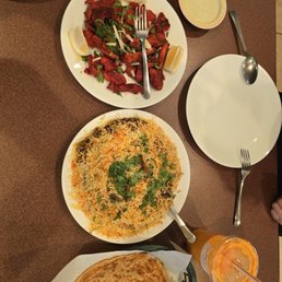 44. Lamb Biryani, Chicken 34. Tandoori Chicken, 54. Paratha - A table features a plate of biryani, a dish of grilled meat with lemon wedges, naan bread, and beverages including a juice and a sauce.