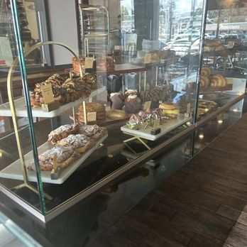 Sucre Table - Baked pastries - A bakery display case contains an assortment of baked pastries, including croissants, cakes, and other desserts. The case is made of glass with a metal frame, and the baked goods are arranged on white trays with small labels. The bakery interior features a modern design with large windows showing parked cars and street activity outside. The reflection on the glass indicates a bright, daytime setting. - Bakeries Near Me - Tampa, Florida