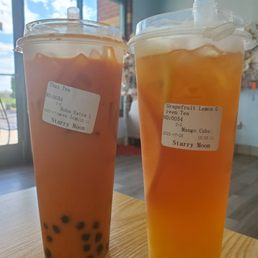Lemon Green Tea - Two large bubble tea drinks in clear plastic cups with lids and straws are placed on a wooden table inside a cafe. The left drink contains a pinkish tea with black tapioca pearls at the bottom, labeled with a sticker indicating Thai tea. The right drink contains an orange-colored tea with visible slices of fruit, labeled as grapefruit lemon green tea with mango. The background shows a window with a view of the sky and part of the cafe interior.