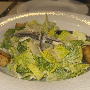 Camille's - Ceasar salad - A Caesar salad featuring chopped romaine lettuce, anchovies placed on top, and grated cheese, served on a white, textured ceramic plate. The setting appears to be in a restaurant with a dimly lit ambiance, and a person is partially visible in the background. - Italian Near Me - Providence, Rhode Island