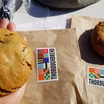 ThoroughBread - Huge $4 cookies. Pb&j (L) & banana Nutella (R) - favorite - The image shows two cookies on brown paper bags with stickers from ThoroughBread, a bakery in Austin, TX. The larger cookie is being held by a hand, while the smaller cookie rests on the paper. The cookies are golden-brown and appear freshly baked. The background includes a white surface and some partially visible items, indicating an outdoor or casual setting. - Bakeries Near Me - Austin, Texas