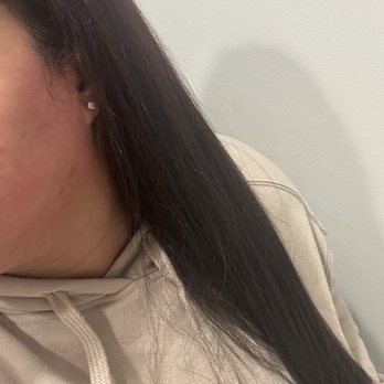 Ars Threading Salon - The end result of the cut/trim and brief blowout. - Close-up image of a woman with dark hair, wearing a beige hoodie and a small earring, against a plain white wall. - Threading Services Near Me - Ozone Park, New York