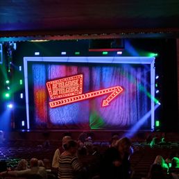 The image shows a theater stage with a neon sign displaying "Betelgeuse" and an arrow, surrounded by colorful stage lighting and an audience seated in front.