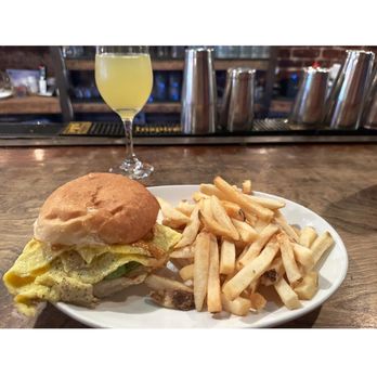 Skytown - A plate containing a sandwich with a bun, egg, and vegetables, alongside a portion of French fries, is positioned on a wooden bar counter. Behind the counter, there is a glass with a yellow beverage and bar equipment including metal shakers and a rubber bar mat. The setting appears to be a bar or casual dining establishment. - New American Near Me - Brooklyn, New York