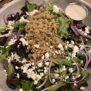 Logan’s Table - Power salad - A salad featuring a bed of mixed greens, including lettuce and arugula, topped with cooked quinoa, crumbled feta cheese, sliced red onions, and roasted eggplant pieces. A small metal cup of creamy dressing is placed on the side. The salad is served on a metal tray, with a wooden table surface visible in the background. - New American Near Me - Leominster, Massachusetts