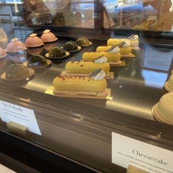 Sucré  - Bakery foods - so yummy - A glass display case contains an assortment of pastries, including pink dome-shaped desserts, green glazed confections, and yellow elongated cakes with toppings. The pastries are arranged in neat rows, with some labeled as 'Nid d'Abeille' and 'Cheesecake.' The reflection of the interior and outdoor scene can be seen on the glass surface. - Desserts Near Me - Covington, Louisiana