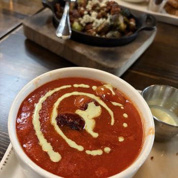Bosscat Kitchen & Libations - GARDEN TOMATO SOUP - A close-up of a bowl of tomato soup with a swirl of cream and a cherry tomato on top. The soup is served in a white bowl placed on a white plate, with a small metal container of cream beside it. The background shows a wooden table with other dishes and utensils, indicating a restaurant setting. - Bars Near Me - Orange, California