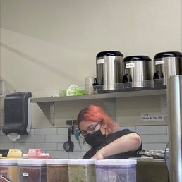 Making the magic mix of the bobas. - A woman with red hair, glasses, and a black mask is working behind a counter with various containers and equipment, in a cafe or tea shop setting.