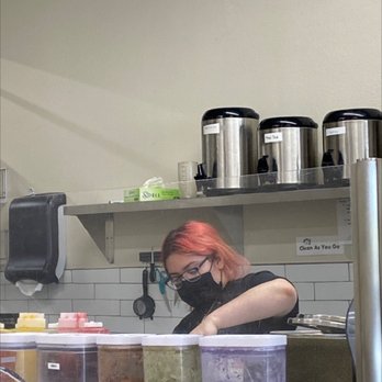 Hi Boba - Making the magic mix of the bobas. - A woman with red hair, glasses, and a black mask is working behind a counter with various containers and equipment, in a cafe or tea shop setting. - Coffee & Tea Near Me - Rio Rancho, New Mexico