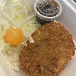 Curry Croquette - A breaded fried cutlet, likely pork or chicken, is presented on a white tray alongside shredded cabbage and a small plastic container of dipping sauce. The cabbage includes a decorative flower-shaped carrot slice. The meal appears to be served in a casual dining setting, possibly a Japanese or Asian-inspired eatery.