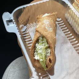 Pistachio Cannoli - A pistachio cannoli with a crispy shell and green pistachio filling, topped with white crumbles, in a clear plastic container.