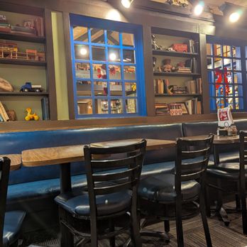 Arni's Restaurant - Old school decor - The image shows a restaurant interior with a long wooden table and black chairs with padded seats. Behind the table, there are bookshelves filled with books, toys, and decorative items. The wall has three blue window frames, with the middle one showing a view into another room decorated with holiday ornaments. The ceiling has exposed beams and multiple ceiling lights, contributing to a warm ambiance. - Pizza Near Me - Lafayette, Indiana