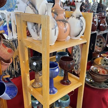 Rail Yards Market - A market stall features a wooden shelf displaying ceramic vases of various sizes and colors, including white with black marbling, peach, and white with black accents. Below, there are ceramic goblets in different colors such as black, blue, red, and green. The background shows shoppers and other market stalls, indicating a busy indoor market environment. - Farmers Market Near Me - Albuquerque, New Mexico