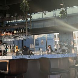Bar area - A bar area with a marble countertop, bar stools, and shelves of liquor bottles, illuminated by sunlight through large windows.