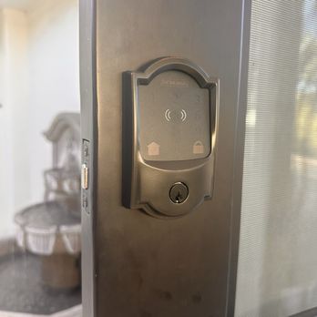 Desert Locksmith - A close-up of a metallic keypad lock with a fingerprint scanner mounted on a door frame. The lock has icons indicating lock and unlock functions, and a keyhole at the bottom. The background shows part of a room with a chair and a basket, slightly out of focus. - Keys & Locksmiths Near Me