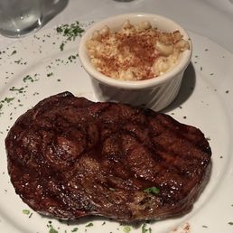RIBEYE, Truffled Bacon Mac & Cheese - A ribeye steak with grill marks is plated with a side of macaroni and cheese in a white dish, garnished with chopped herbs.