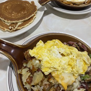 Alexander's Restaurant - The image shows a breakfast setting with two brown ceramic dishes containing scrambled eggs on top of hash browns and potatoes, and a fried egg in a separate dish. There are also two plates with pancakes visible in the background. The dishes are placed on a white marble or laminate table surface. - Diners Near Me - Chicago, Illinois