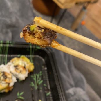 Sushi D - Dragon Roll - A piece of sushi with fish, toppings, and sauce is held by chopsticks in the foreground. The sushi tray below contains more sushi pieces with similar toppings. The background includes a wooden table and a dark surface, indicating a dining setting, likely at a Japanese restaurant or sushi bar. - Sushi Bars Near Me - Brooklyn, New York