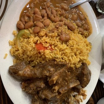 Rico Pollo - Hot line dish - A white oval plate contains yellow rice with vegetables, a portion of beans in sauce, and a large piece of cooked meat, possibly beef or goat, with bones. A metal spoon rests on the edge of the plate. The dish appears to be a meal served at a restaurant or home setting. - Spanish Near Me - Brooklyn, New York
