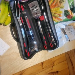 Grill set from Walmart - A barbecue tool set containing utensils such as tongs, a spatula, and a basting brush is arranged inside a black carrying case with elastic straps. The case is open on a wooden table, with grocery bags and bananas nearby, indicating a shopping or preparation scene.