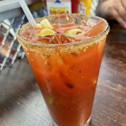 Bloody Mary - A Bloody Mary cocktail is served in a tall glass with a salted rim. The drink contains tomato juice and is garnished with vegetables, including pickles and possibly olives. The glass is placed on a wooden table, with condiments like ketchup and mustard visible in the background. A straw is inserted into the drink, and part of a person's arm is visible in the background.