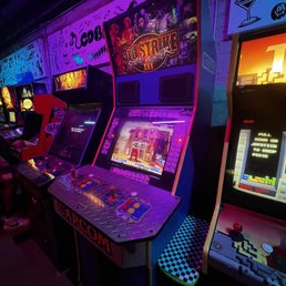 Arcade games - A row of vintage arcade game machines, including Street Fighter, are positioned against a wall in a dimly lit arcade bar. The machines have colorful screens and controls, with some featuring artwork and branding. The environment includes wall decorations with playful designs and lighting that emphasizes the gaming area. The arcade setup suggests a lively, entertainment-focused space for gaming enthusiasts.