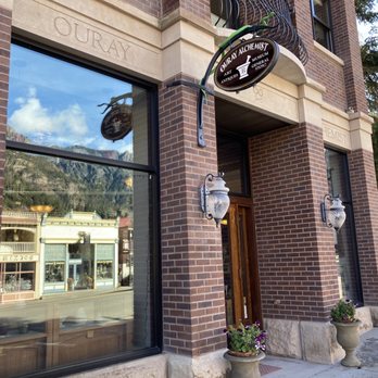 Ouray Alchemist - The exterior of the building. - The photo depicts the brick facade of Ouray Alchemist museum, with a sign hanging above the entrance indicating it is a museum and general store. Large windows reflect the street scene and mountains in the background. The entrance is flanked by two decorative outdoor lamps, and there are flower pots with plants near the door. The building has stone accents and engraved names, with a sidewalk in front. - Museums Near Me - Ouray, Colorado