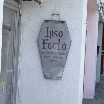 Ipso Facto - Sign - A metal sign with purple lettering displays 'Ipso Facto' and describes the store's offerings, including dark apparel, accessories, and esoteric items, mounted on a white exterior wall. - Shoe Stores Near Me - Fullerton, California