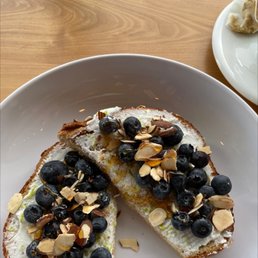 Blueberry Ricotta Toast - A slice of blueberry ricotta toast topped with fresh blueberries and sliced almonds, served on a white plate. The toast appears to have a layer of ricotta cheese and is garnished with blueberries and almond slices, with a drizzle of honey or syrup. The background shows a wooden table and part of another plate with food.