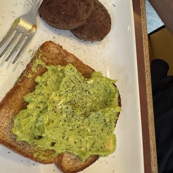 105 Cafe - Avocado toast - A breakfast meal is presented on a white rectangular plate, including two cooked sausage patties, a slice of toasted bread topped with mashed avocado, and sprinkled with black pepper. A fork rests on the plate, and part of a person's arm is visible in the upper right corner. The plate is placed on a wooden table, and the setting appears to be a casual dining environment. - American Near Me - Conroe, Texas
