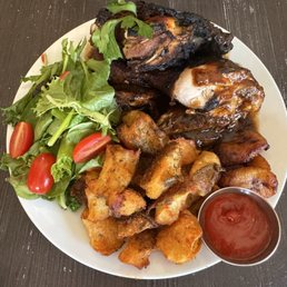 A plate with grilled meat, fried potatoes, a side salad with cherry tomatoes, and a small cup of ketchup on a dark wooden table.