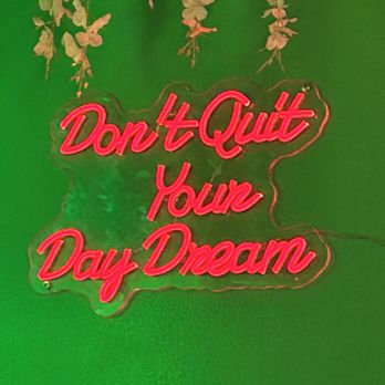 Casa Maya - A neon sign with bright pink cursive lettering states 'Don't Quit Your Day Dream' and is mounted on a green wall. Above the sign, there are hanging decorative elements resembling dried or artificial flowers or leaves. The sign appears to be made of glass or acrylic with neon tubing, and the overall setting suggests an interior space with decorative lighting. - Bars Near Me - Brooklyn, New York