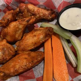 Buffalo chicken wings with celery and carrot sticks, accompanied by a cup of ranch dressing, served on checkered paper.