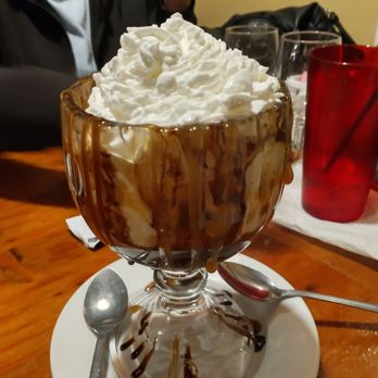 Bubb's Grub - Delicious chocolate cake sundae! - A tall glass sundae with whipped cream and chocolate syrup, served on a white plate with a spoon, on a wooden table. - Barbeque Near Me - Catalina, Arizona