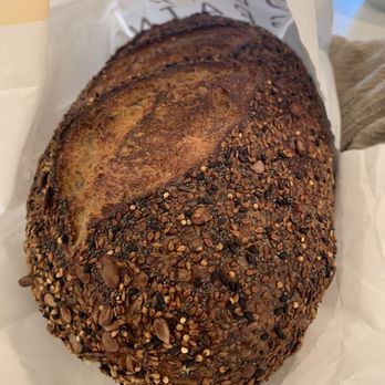 Dear Grain - a loaf of bread - A loaf of bread with a golden-brown crust, topped with a mixture of seeds including sesame, sunflower, and flax seeds. The bread appears freshly baked, with a textured surface and a slightly cracked crust. It is partially wrapped in white paper with some printed text visible, placed on a surface with a cloth and a cup nearby. - Bakeries Near Me - Hamilton, Ontario