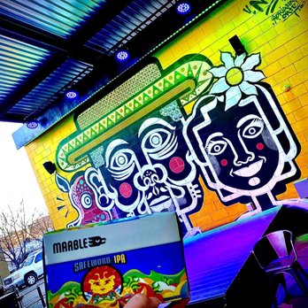 Marble Brewery - A mural with stylized, cartoon-like faces with bold outlines and bright colors is painted on a yellow brick wall at Marble Brewery in Albuquerque, NM. The mural depicts multiple faces with expressive features, including large eyes and red cheeks. The scene is illuminated by colorful lighting from above, and outdoor elements such as parked cars and leafless trees are visible in the background. A person holds a beer coaster or card in the foreground, partially obscuring the lower part of the mural. - Breweries Near Me - Albuquerque, New Mexico
