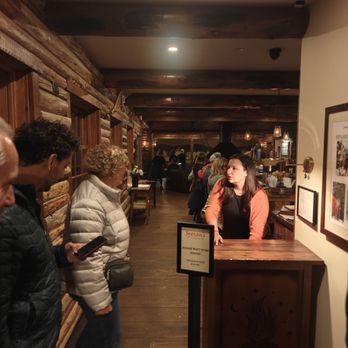 Johnny Seesaw's - A woman stands behind a reception desk in a rustic restaurant with wooden walls and ceiling beams, while guests wait in line. - American Near Me - Peru, Vermont