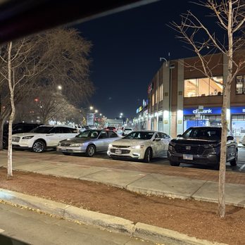 Veggie House - Complex - The image shows a parking lot at night with several parked cars, including sedans and SUVs, in front of a shopping center. Leafless trees line the sidewalk, and the storefronts are lit up, indicating commercial activity. The sky is dark, and streetlights illuminate the area, with some buildings visible in the background. - Vegan Near Me - Chicago, Illinois