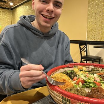 Ghar- E- Kabab - Ethiopian vegetable Veggi Combo 7 Type Shro,misir,kiki,defin Misir,dinch,cabbage - A young man with a shaved head and a gray hoodie is smiling at the camera, holding a fork near a large salad bowl filled with various vegetables. - Indian Near Me - Silver Spring, Maryland