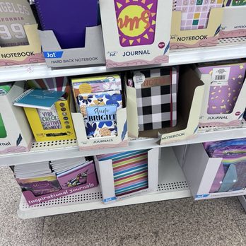 Dollar Tree - The image shows retail store shelves stocked with a variety of notebooks and journals. The top shelf features boxed journals with bright, patterned covers and motivational phrases like 'Shine Bright.' The middle shelf contains more notebooks, including a black-and-white checkered design and others with floral and striped patterns. The bottom shelf displays smaller notebooks and journals, some with inspirational themes. The store floor is visible with a speckled tile surface. - Discount Store Near Me - Pinole, California
