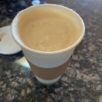SoPo Coffee - Cafe Misto - A disposable paper cup filled with a cafe misto, featuring a layer of frothy milk on top, is placed on a dark, textured countertop. The cup has a cardboard sleeve for insulation, and a plastic lid is visible nearby. The background includes a blurred metallic object and a small white item, indicating a casual coffee shop setting. - Coffee & Tea Near Me - Albuquerque, New Mexico