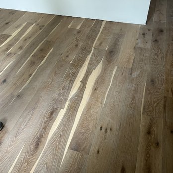 Ideal Design Center - The floor I got - The image shows a section of an interior room with wood flooring composed of wide planks. The flooring has a natural wood finish, displaying visible grain patterns, knots, and color variations from light to darker shades. A white wall is visible at the top of the image, and a person's foot wearing a black shoe is partially seen in the lower left corner. The flooring appears to be newly installed or well-maintained. - Flooring Near Me - Hyannis, Massachusetts
