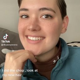 Pixie hair cut - A person with short, dark hair and light skin is smiling at the camera, wearing a teal zip-up jacket and a brown shirt underneath. The background shows a wall with a painting.