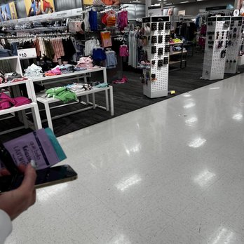 Target - The image shows a retail store section dedicated to children's clothing and accessories. Clothing items such as socks, shirts, and shorts are arranged on tables and racks. Promotional posters featuring children are visible above the displays. The store has bright lighting, a black carpeted area for the clothing displays, and a white tiled floor in the foreground. A person is holding a phone and a shopping list in the lower left corner of the image. - Department Stores Near Me - Cincinnati, Ohio
