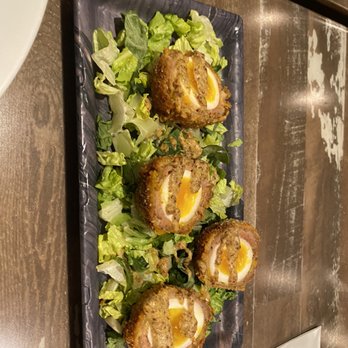 Jackdaw - Scottish eggs - Four Scotch eggs, each halved to reveal the cooked yolk inside, are placed on a black rectangular plate. The eggs are breaded and fried, with a crispy exterior. They are served on a bed of chopped lettuce leaves, which add a fresh contrast to the dish. The presentation is simple and focused on the food, with a rustic wooden table surface visible in the background. - Irish Near Me - Fort Worth, Texas